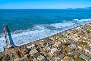 An aerial view of a coastal residential area with houses, a pier extending into the sea, and a clear sky.