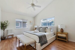 Quarto com cama, mesas de cabeceira, um banco, ventilador de teto e janelas.