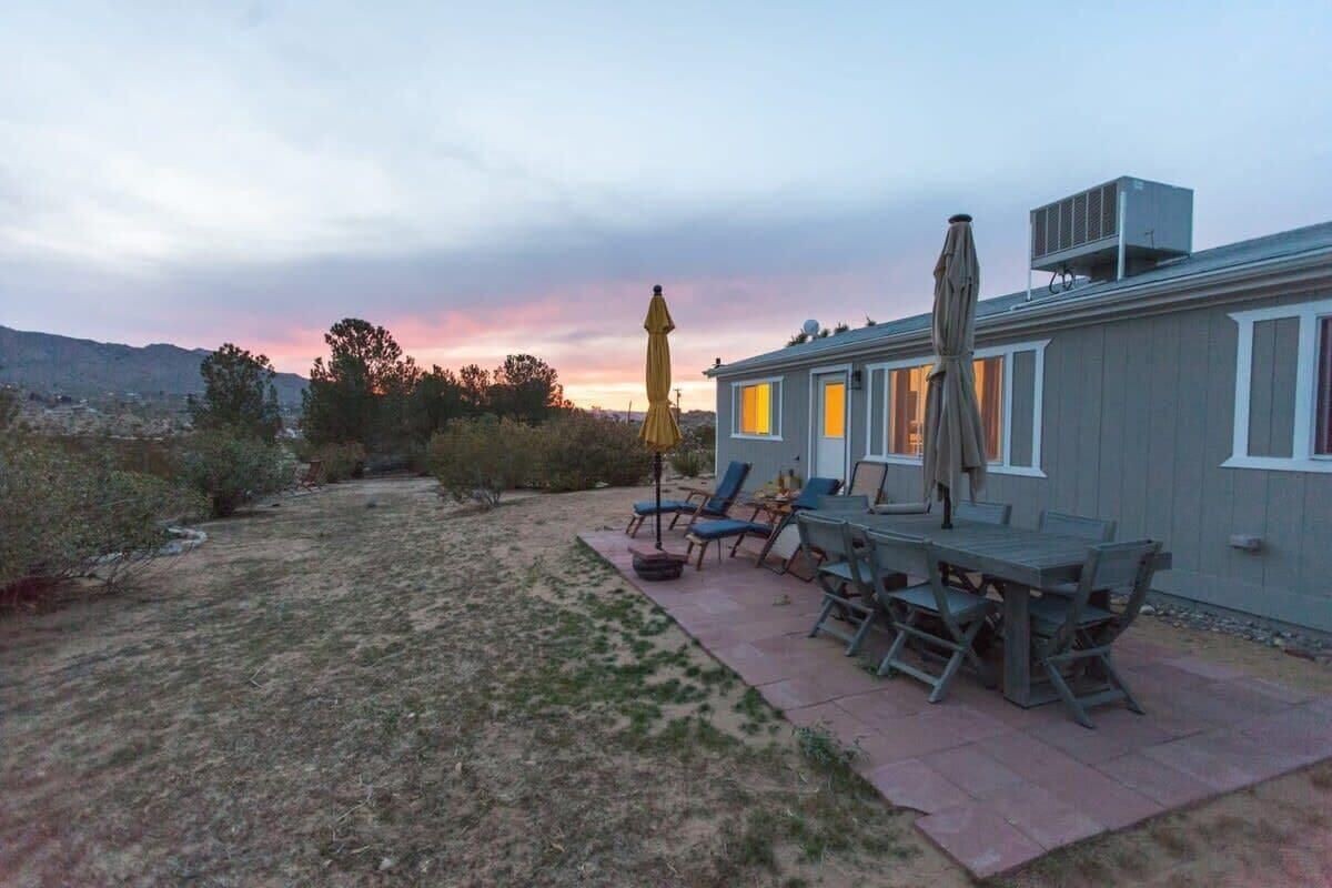 A patio with outdoor furniture and a yellow umbrella, a house with a gray exterior, and a sunset in the background.