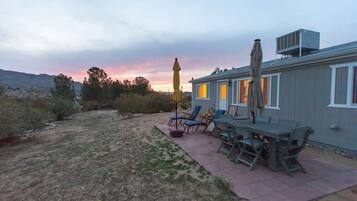 A patio with outdoor furniture and a yellow umbrella, a house with a gray exterior, and a sunset in the background.