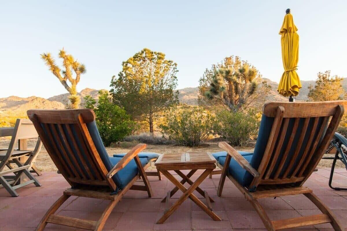 A wooden table and two chairs with blue cushions, set outdoors with a view of desert vegetation and a mountain in the background.