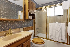 Un baño con tocador de madera, inodoro blanco, ducha con cerramiento de vidrio y espejo con diseño de papel tapiz floral.