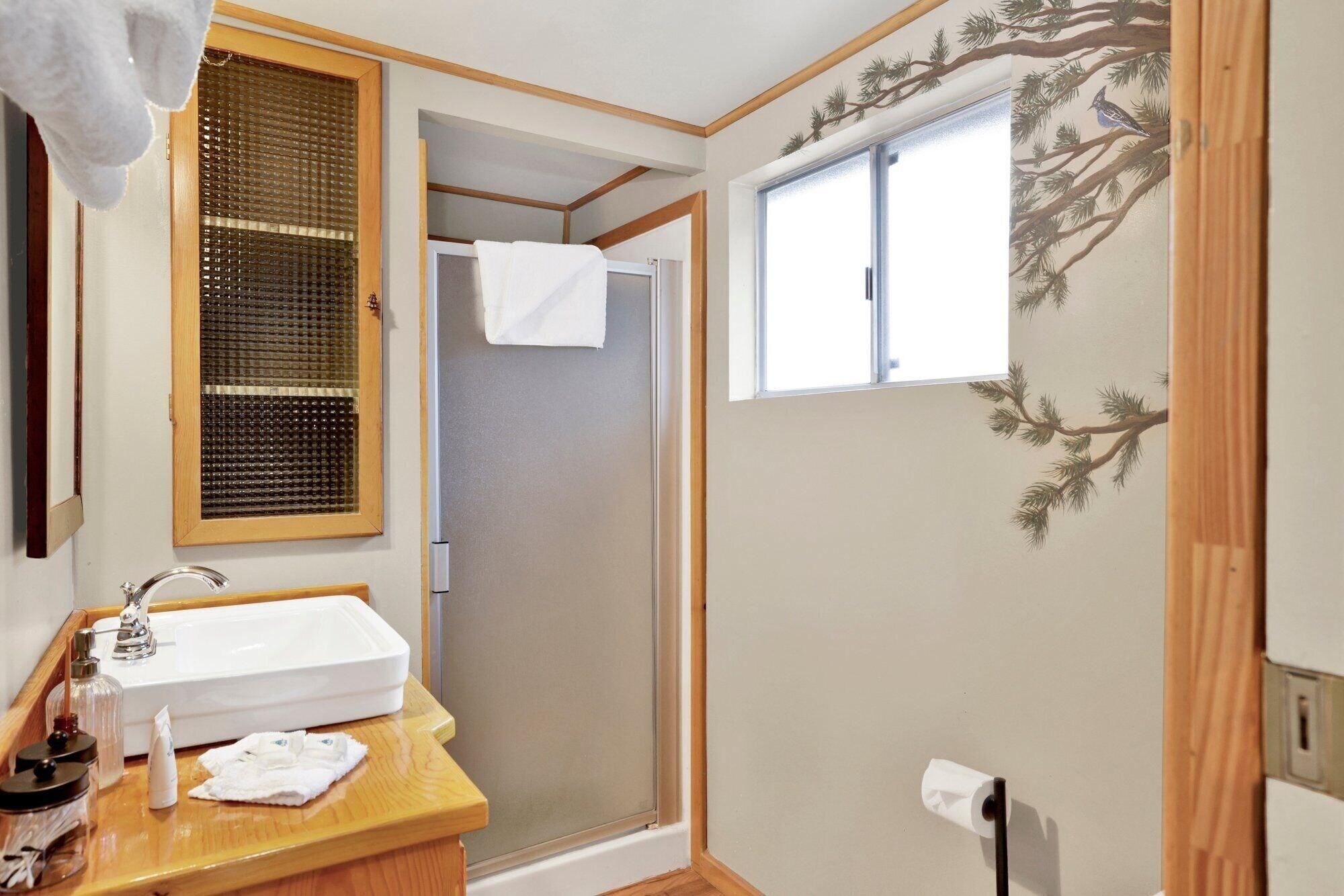 A bathroom with a wooden vanity, a white sink, a towel, a window with a tree mural, and a shower area.