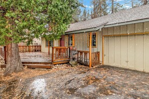 A wooden deck leading to a building with a garage, surrounded by trees and a leaf-covered ground. - Timber Pines Chalet #2417 (Big Bear Lake)