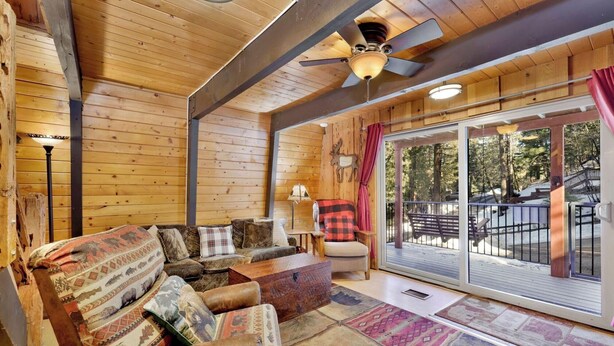 A cozy cabin interior with a wooden ceiling, a ceiling fan, a sofa with patterned cushions, a chair, and a view of a snowy outdoor deck.
