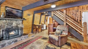 A cozy interior with a stone fireplace, wooden walls, a comfortable armchair, and a staircase.