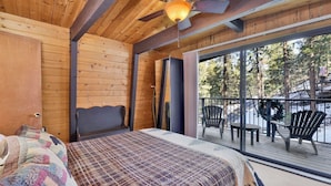 A wooden cabin room with a bed, a ceiling fan, and a balcony with chairs.
