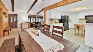 A spacious living area with a wooden dining table, a cozy sofa, and a view through an open doorway to a kitchen with modern appliances. - Casa Bosque #2176 (Big Bear Lake)