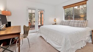 A bedroom with a bed, a desk, a chair, a lamp, and a window with a view of trees. - Casa Bosque #2176 (Big Bear Lake)