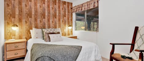 Un dormitorio con cabecera de madera, cama con ropa de cama blanca, mesita de noche con lámpara y ventana con vistas.