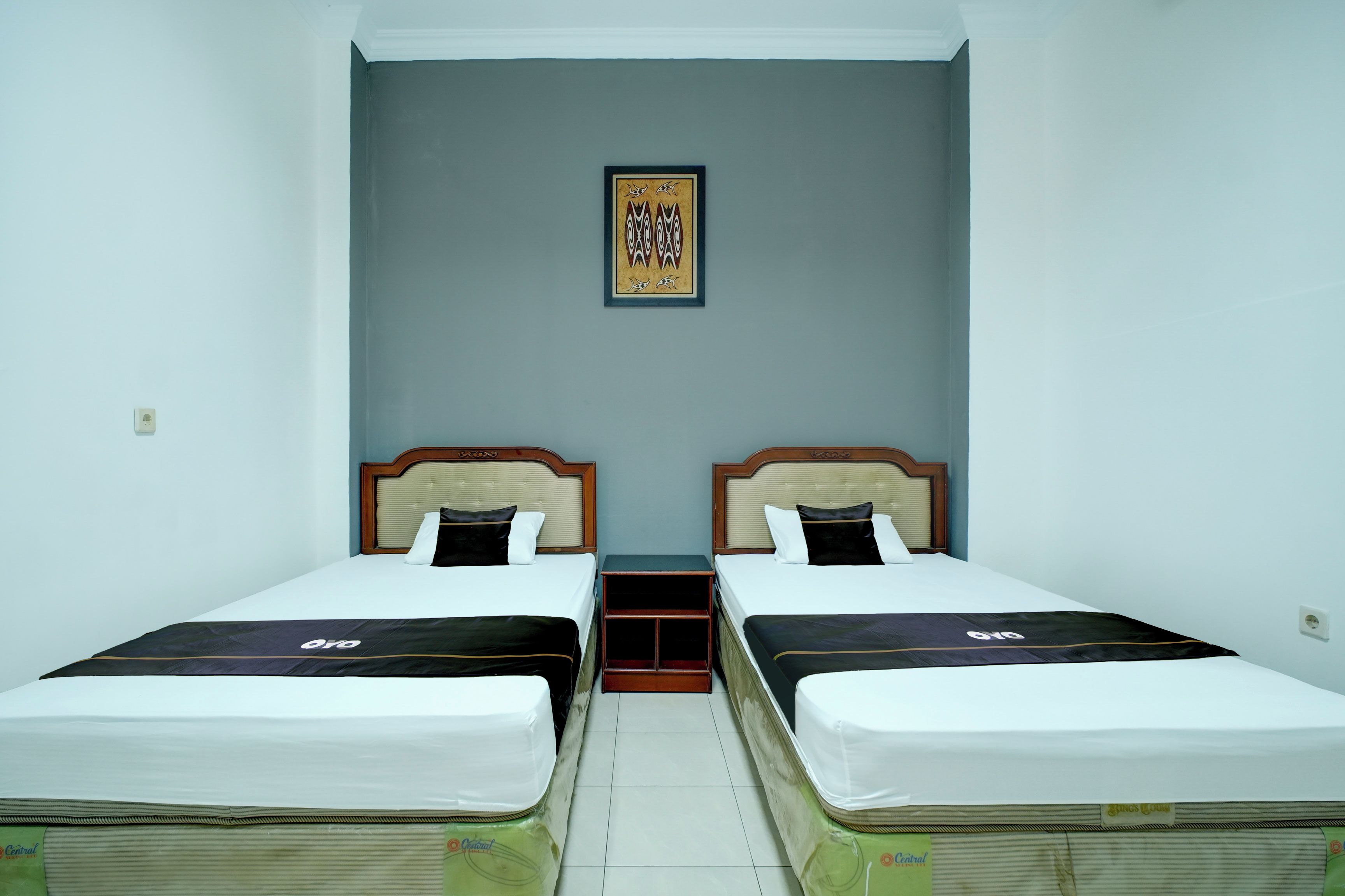 A hotel room with two single beds, a small table, a wall-mounted shelf, and a framed picture.