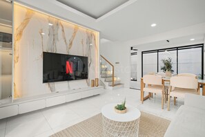 A modern living room with a large flat-screen TV mounted on a marble wall, a dining area with a wooden table and chairs, and a staircase leading to an upper level.
