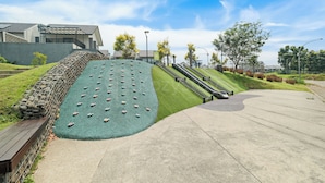 Une aire de jeux avec des structures d’escalade et un toboggan, entourée d’une zone pavée et de verdure.
