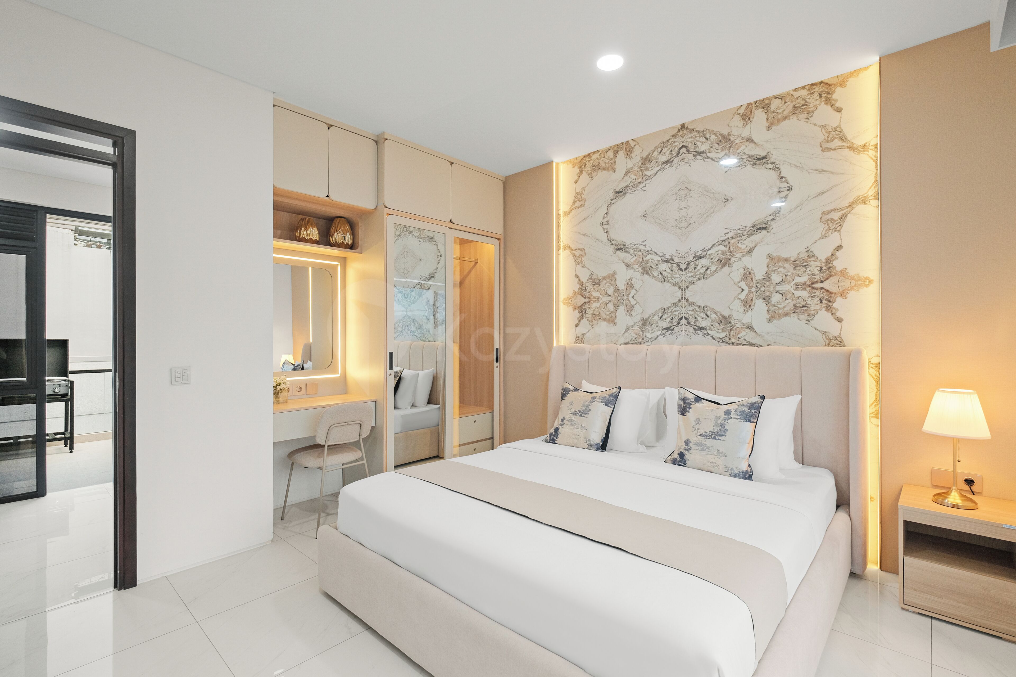A modern bedroom with a large bed, a bedside table, a nightstand, a mirror, a wardrobe, and a bathroom visible through an open door.