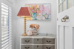 Ein Zimmer mit einer weißen Tür, einem an der Wand hängenden Gemälde, einem Tisch mit einer Lampe und einer Vase mit Blumen.