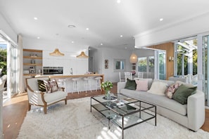A modern living room with a glass coffee table, a sofa with cushions, and a kitchen area with white cabinets and pendant lights.