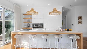 A modern kitchen with a central island, bar stools, pendant lights, and a view through a glass door. - Luxury On Lemon (New Plymouth)