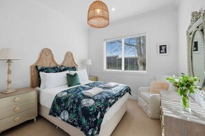 Ein Schlafzimmer mit Bett, Nachttischen, einem Schminktisch, einem Spiegel und einem Fenster mit Jalousien.