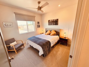 Une chambre à coucher comprenant un lit, une chaise, une table de chevet, un ventilateur de plafond et une fenêtre avec des stores.
