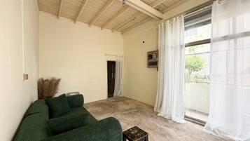 A room with a green sofa, a small stool, a ceiling with wooden beams, a window with white curtains, and a door.