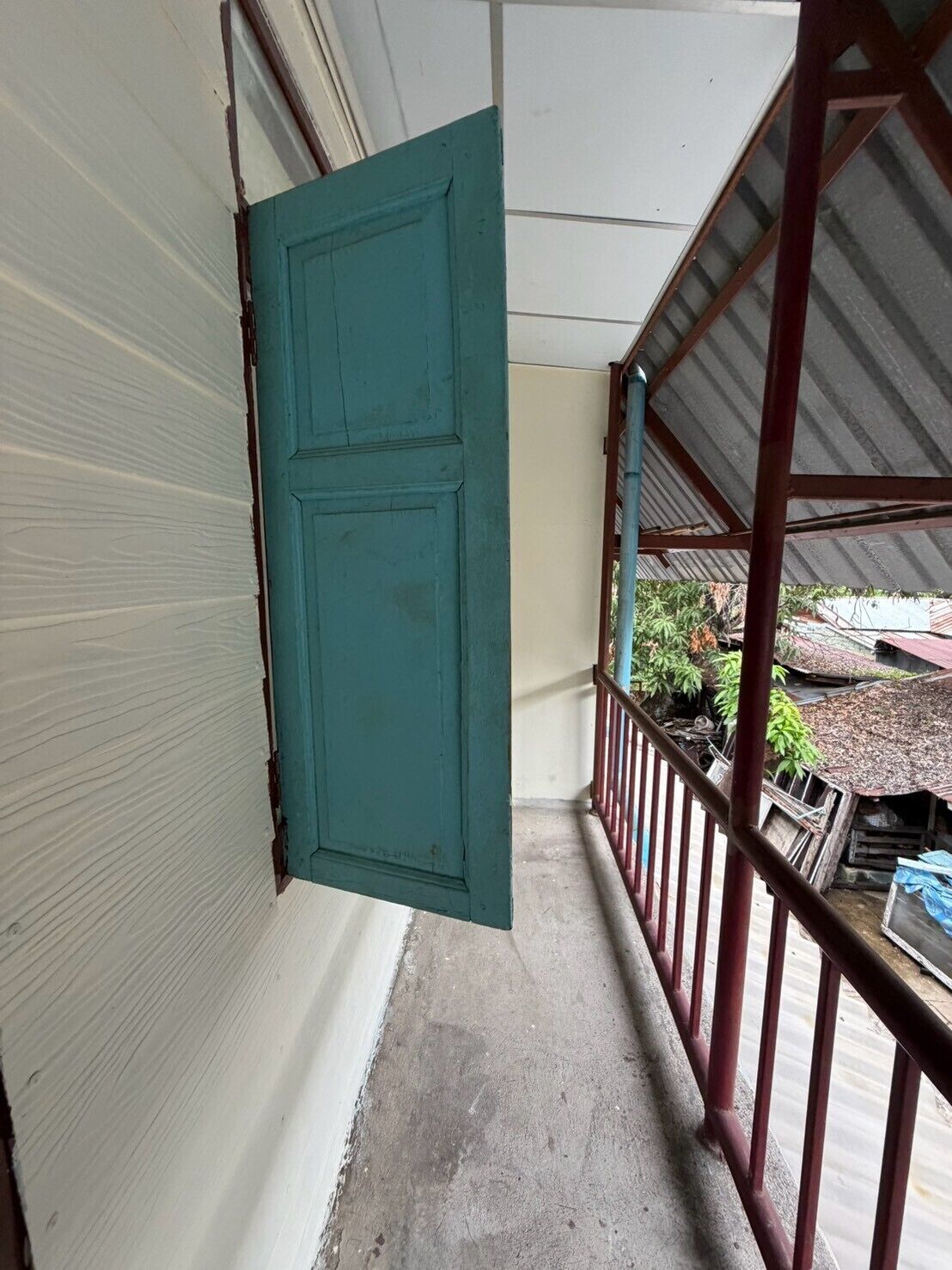 A narrow corridor with a teal door, a metal railing, and a view of a yard with plants and a building in the background.