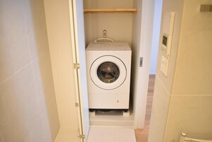 A compact, white laundry machine in a narrow hallway with a wooden shelf above it.