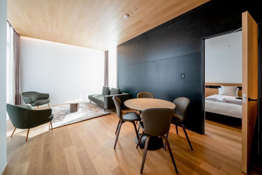 Une chambre d’hôtel moderne avec un plafond en bois, un mur d’accent de couleur foncée, une table à manger ronde avec des chaises, un canapé et un lit doté d’une tête de lit blanche.