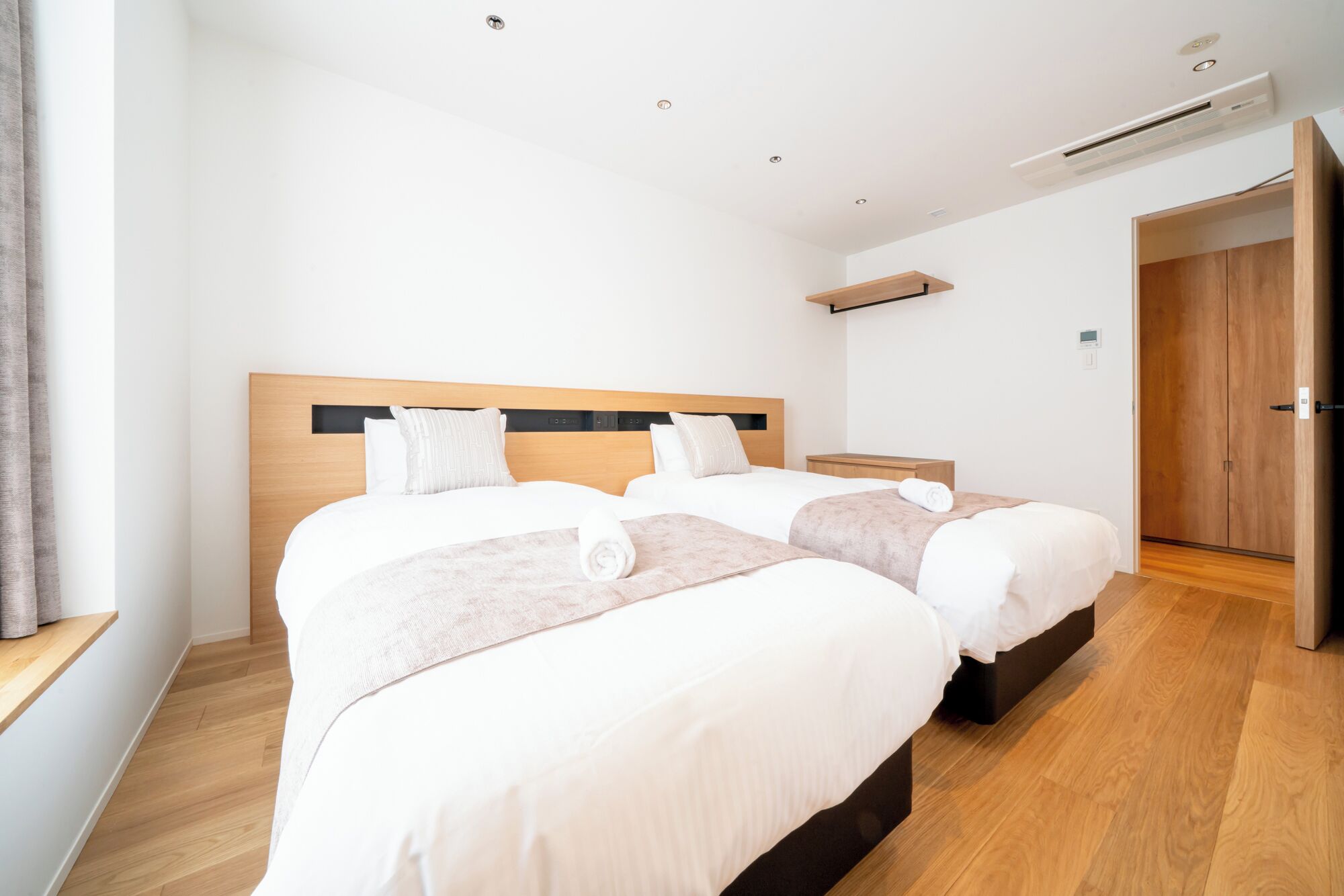 A hotel room with two beds, a wooden headboard, a bedside table, a nightstand, and a door leading to another room.