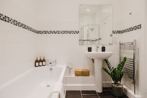 A modern bathroom with a bathtub, a sink, a mirror, and a potted plant. - Stunning Loft Apartment With Private Parking (Chipping Norton)