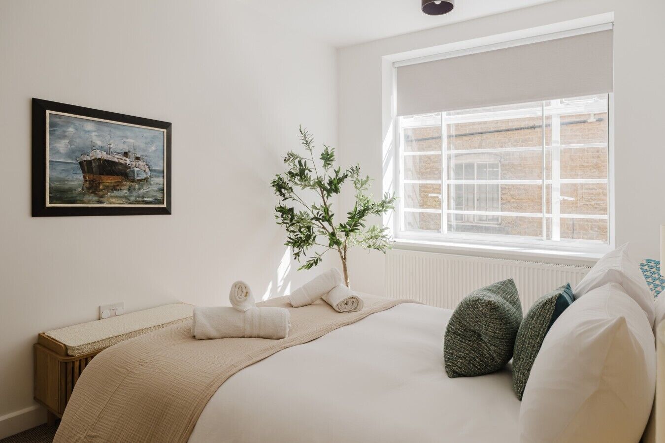 A bedroom with a bed, a window, a radiator, a plant, and a framed picture on the wall.