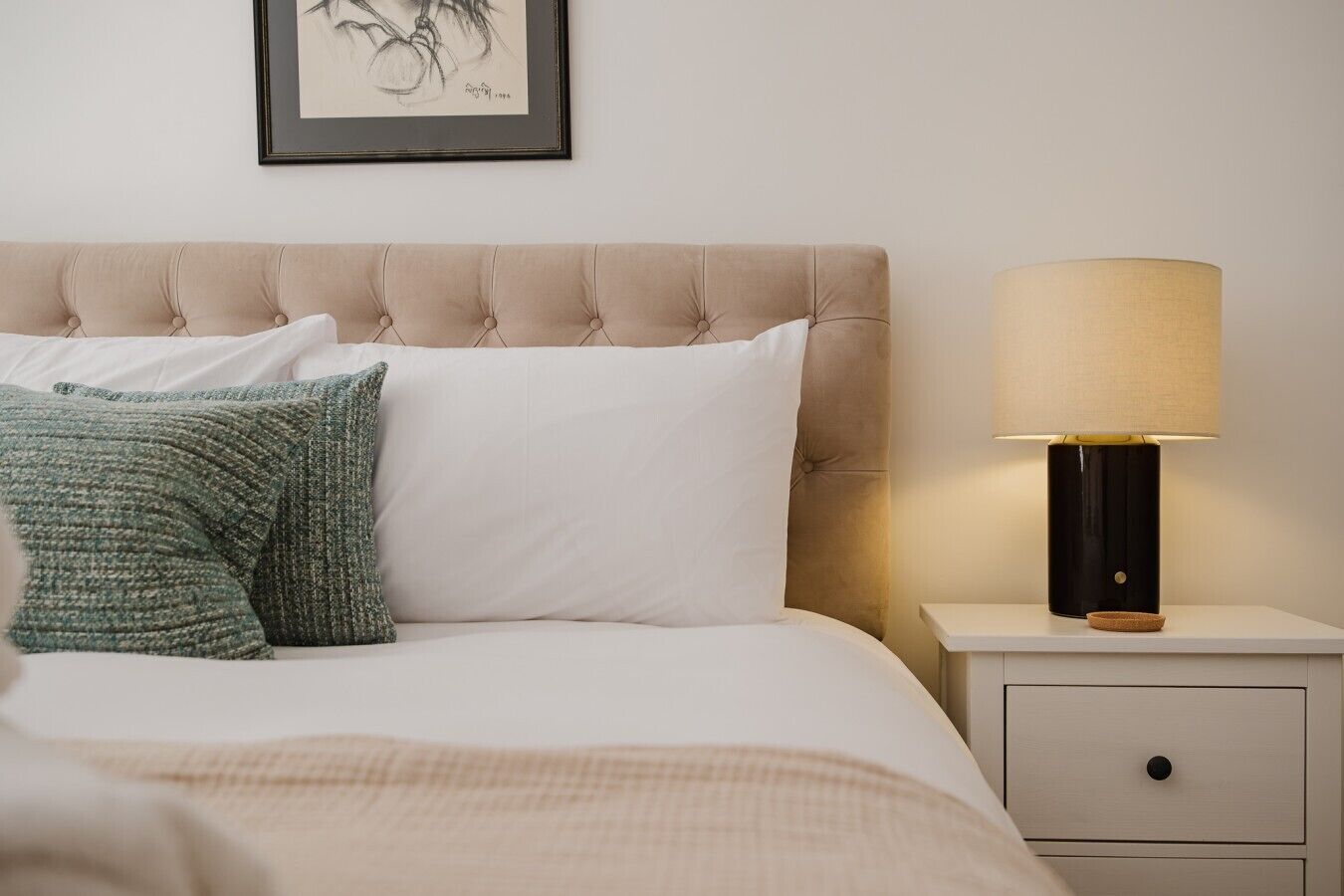 Een keurig opgemaakt bed met een gestoffeerd hoofdbord, een bedlamp en een lijst met een kunstwerk aan de muur.