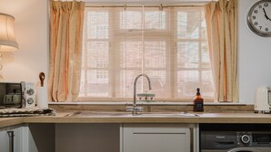 A kitchen with a sink, a washing machine, a clock, and a window with curtains. - Beautiful Cottage in the Heart of Malmesbury (Malmesbury)