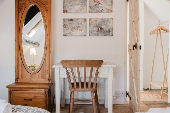 A bedroom with a wooden dresser, a desk with a chair, a mirror, and artwork on the wall.