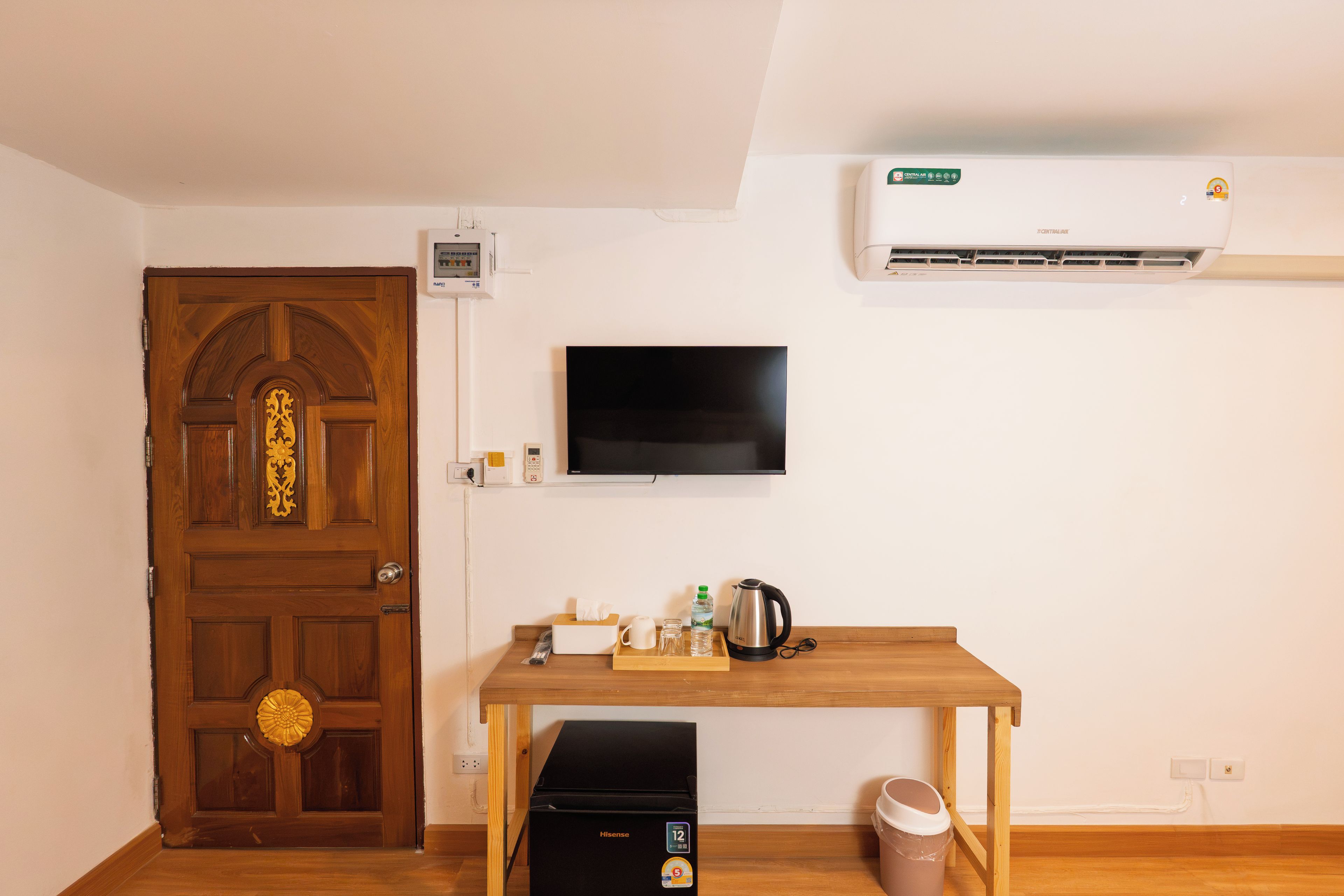 Een kamer met een houten deur, een wandgemonteerde airconditioning, een flatscreen-tv, een tafeltje met een waterkoker, een prullenbak en een waterfles.