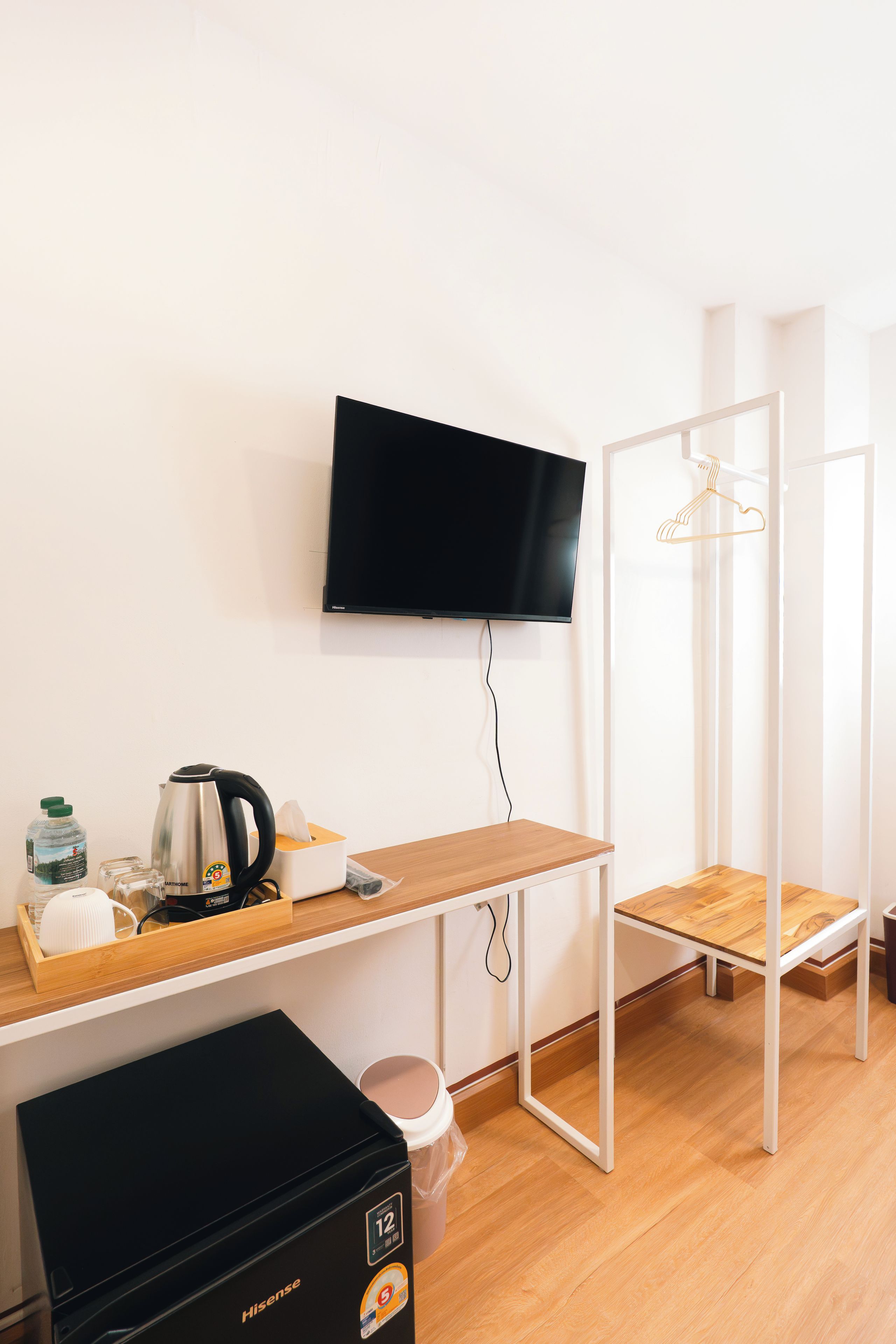 A compact room with a flat-screen TV mounted on the wall, a wooden desk with a kettle, a toaster, and a water bottle. There's a small trash bin and a compact refrigerator.