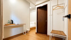 Quarto de hotel com piso de madeira, uma cama, um guarda-roupa, uma mesinha com chaleira e um cabideiro.