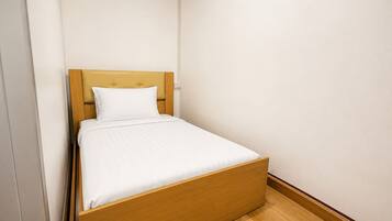 Una cama individual con cabecera de madera y ropa de cama blanca en una habitación con paredes lisas y suelo de madera.
