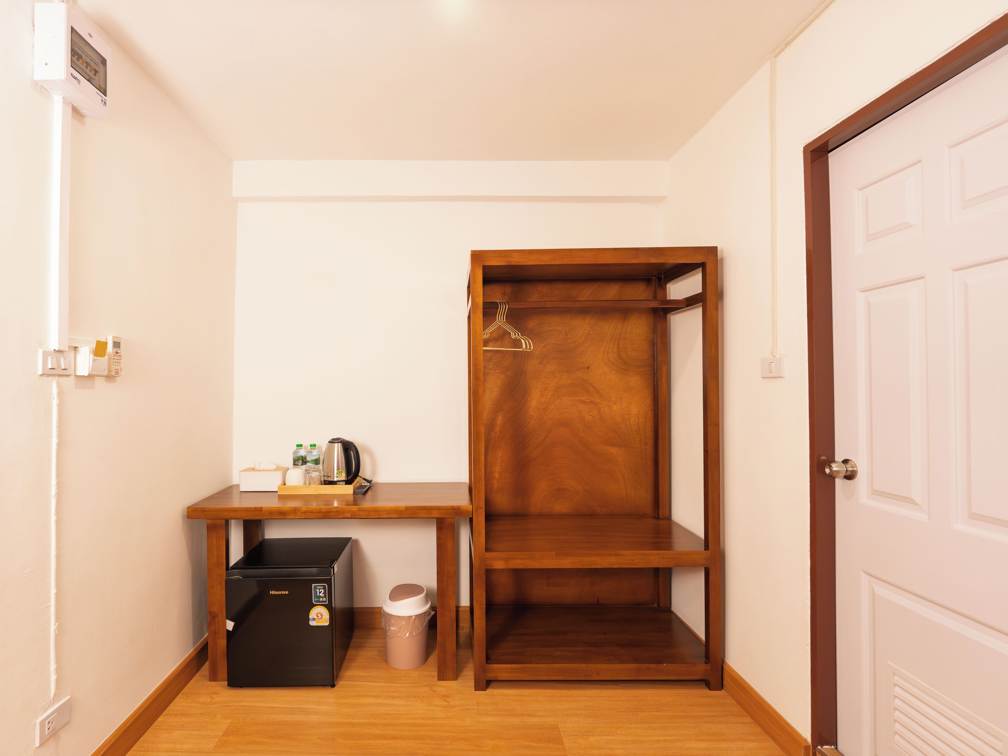 A compact room with a wooden wardrobe, a small table with a kettle, a trash bin, and a compact refrigerator.
