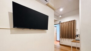 Quarto com televisão montada na parede, piso de madeira, janela com cortinas e uma pequena mesa com chaleira.