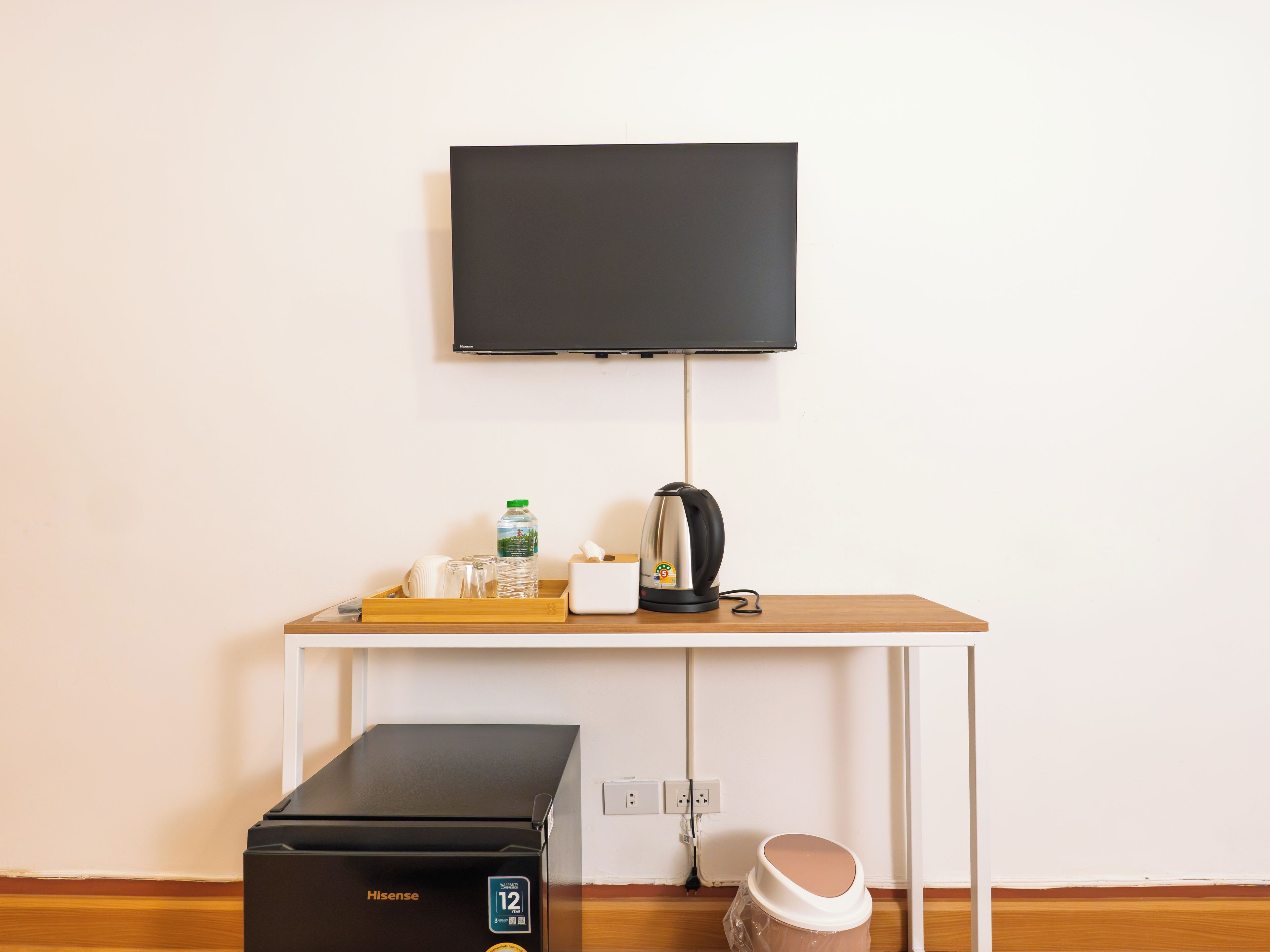 Une chambre dotée d’un téléviseur à écran plat fixé au mur, d’une table en bois avec une bouilloire, une bouteille d’eau et une boîte de mouchoirs, ainsi que d’une imprimante Hisense noire posée sur le sol.