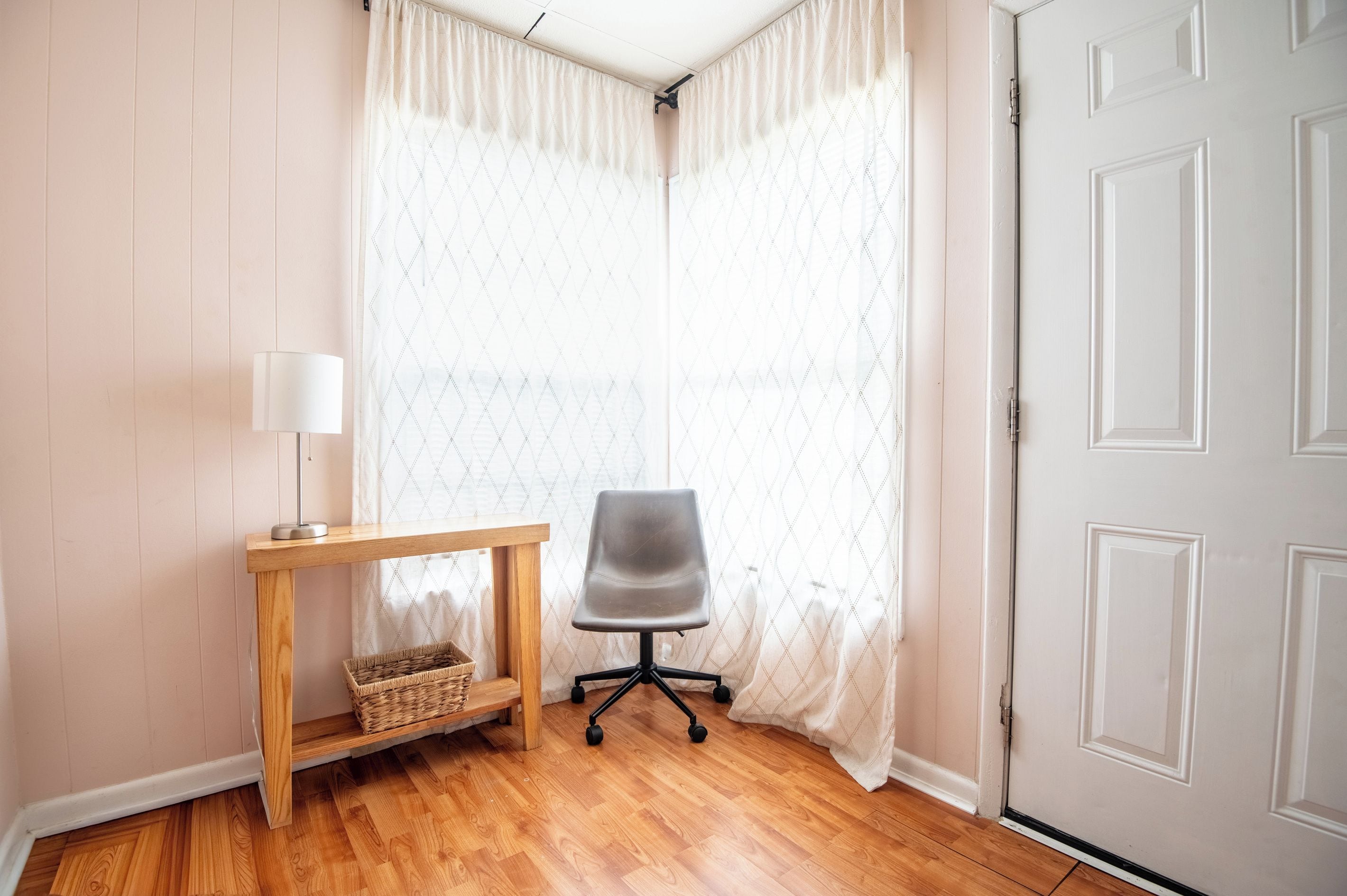 A corner room with a wooden floor, a grey office chair, a small wooden table with a lamp, and a white door.