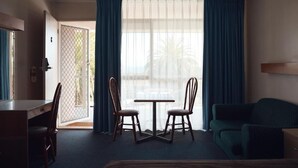 Habitación de hotel con cama, escritorio, dos sillas, una mesa y un sofá. Hay un ventanal con cortinas, y una puerta que da al exterior.