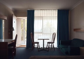 Habitación de hotel con cama, escritorio, dos sillas, una mesa y un sofá. Hay un ventanal con cortinas, y una puerta que da al exterior.