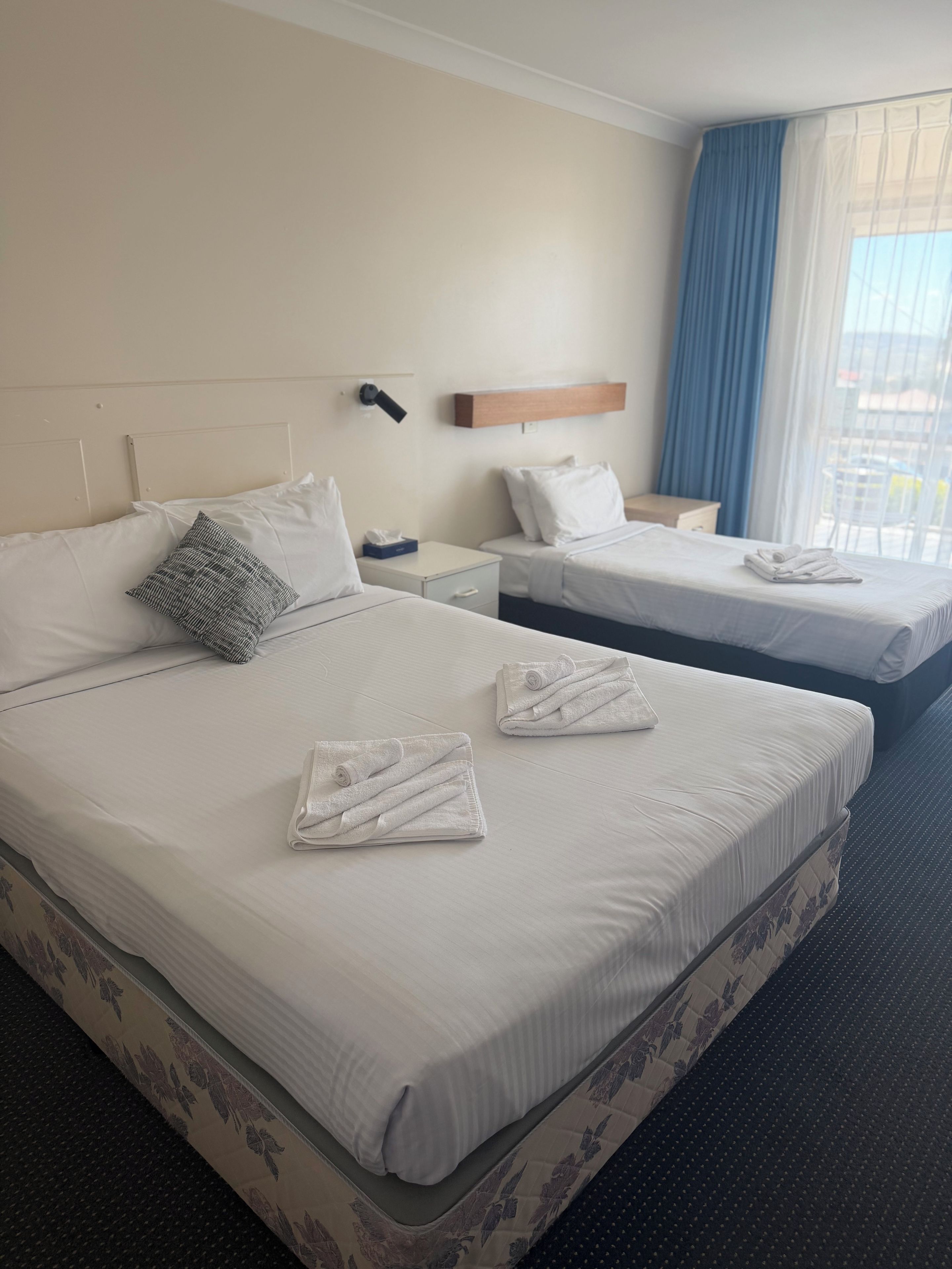 A hotel room with two beds, each with white linens and pillows, a bedside table with a lamp, and a window with blue curtains.