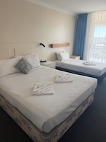 Quarto de hotel com duas camas, cada uma com lençóis e almofadas brancas, mesa de cabeceira com abajur e janela com cortinas azuis.
