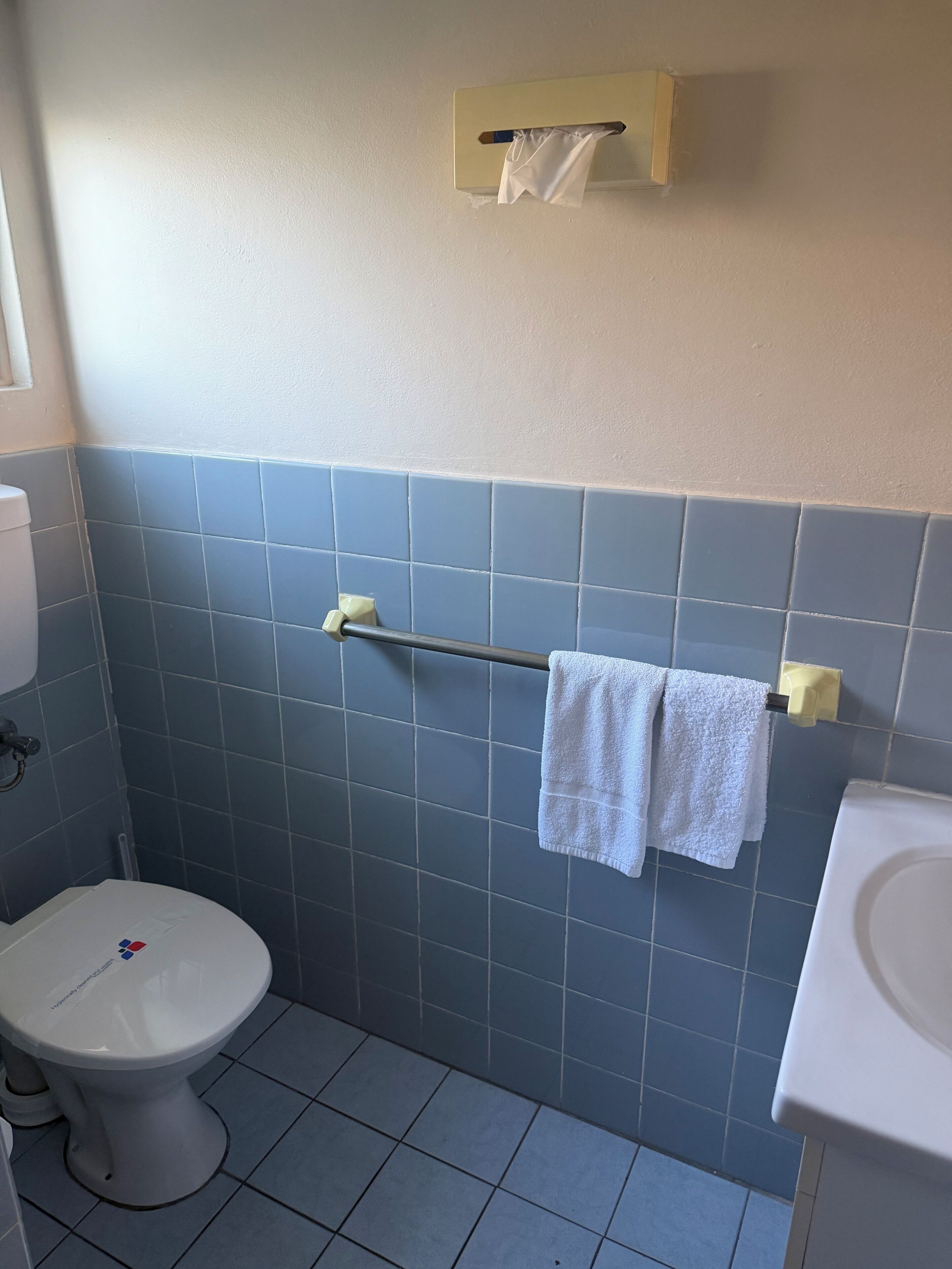 Une salle de bain comprenant des toilettes, un porte-serviettes avec deux serviettes et un distributeur de mouchoirs fixé au mur.