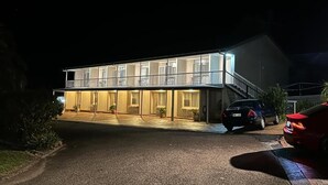Un edificio de dos pisos con balcón, un acceso cubierto y un automóvil estacionado frente a él.