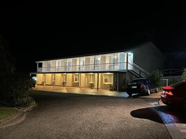 Un edificio de dos pisos con balcón, un acceso cubierto y un automóvil estacionado frente a él.