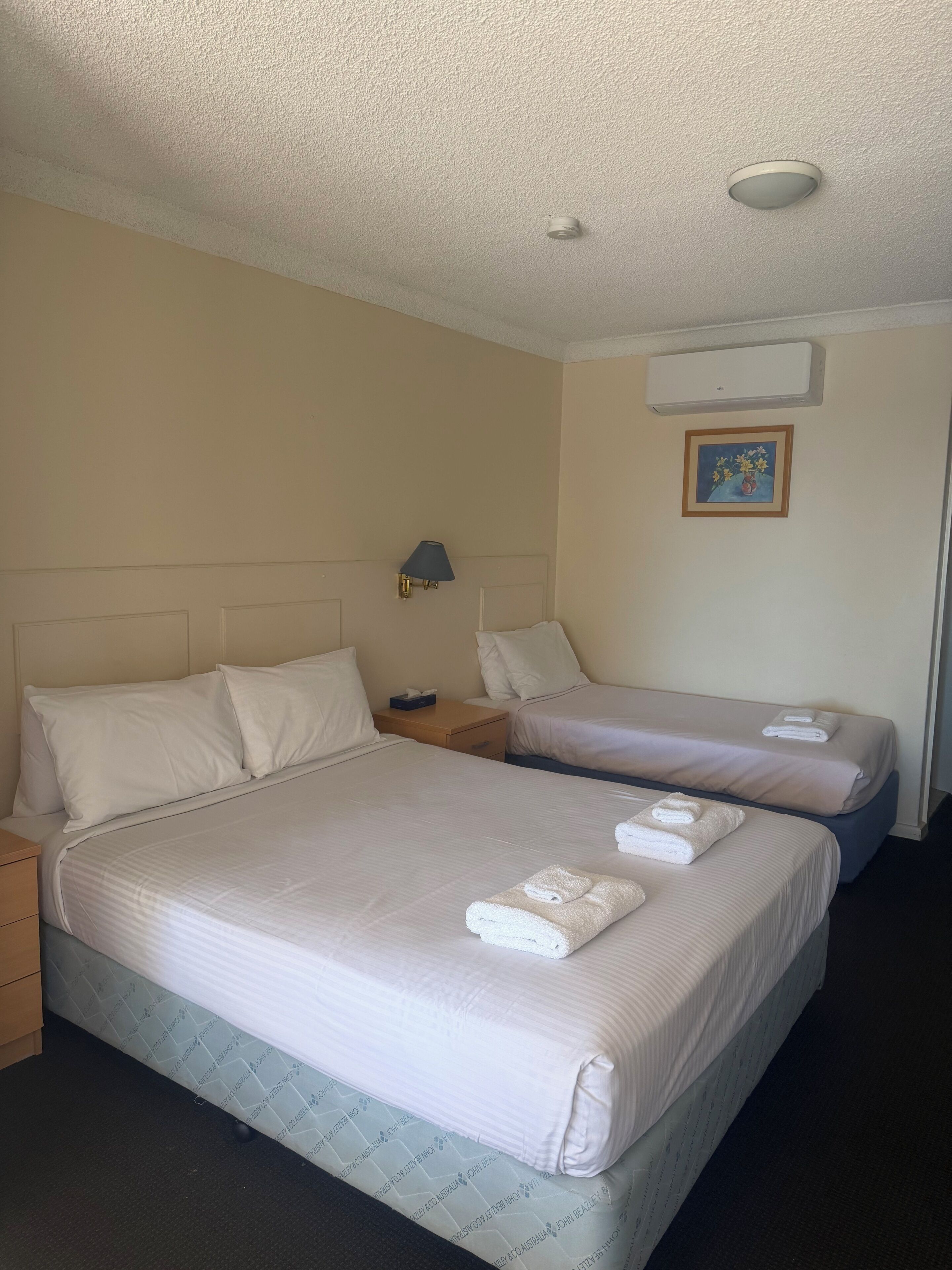 A hotel room with two beds, a nightstand, a lamp, an air conditioning unit, and a framed picture on the wall.