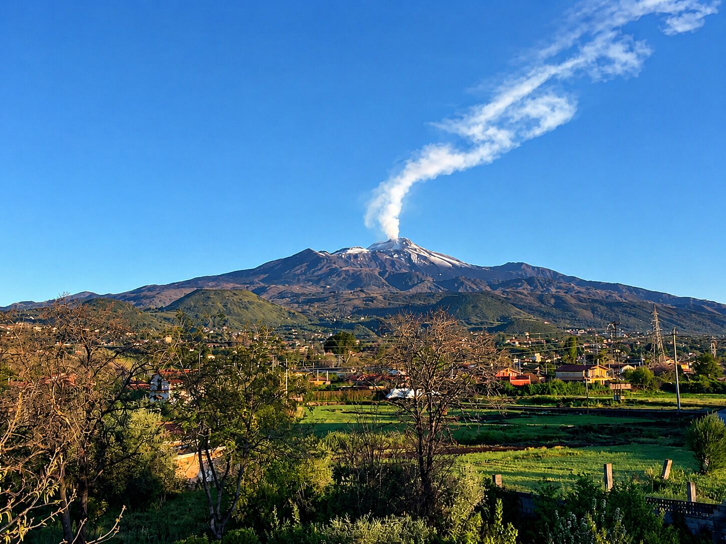 A mountain with a visible smoke plume, surrounded by a rural landscape with houses and greenery.